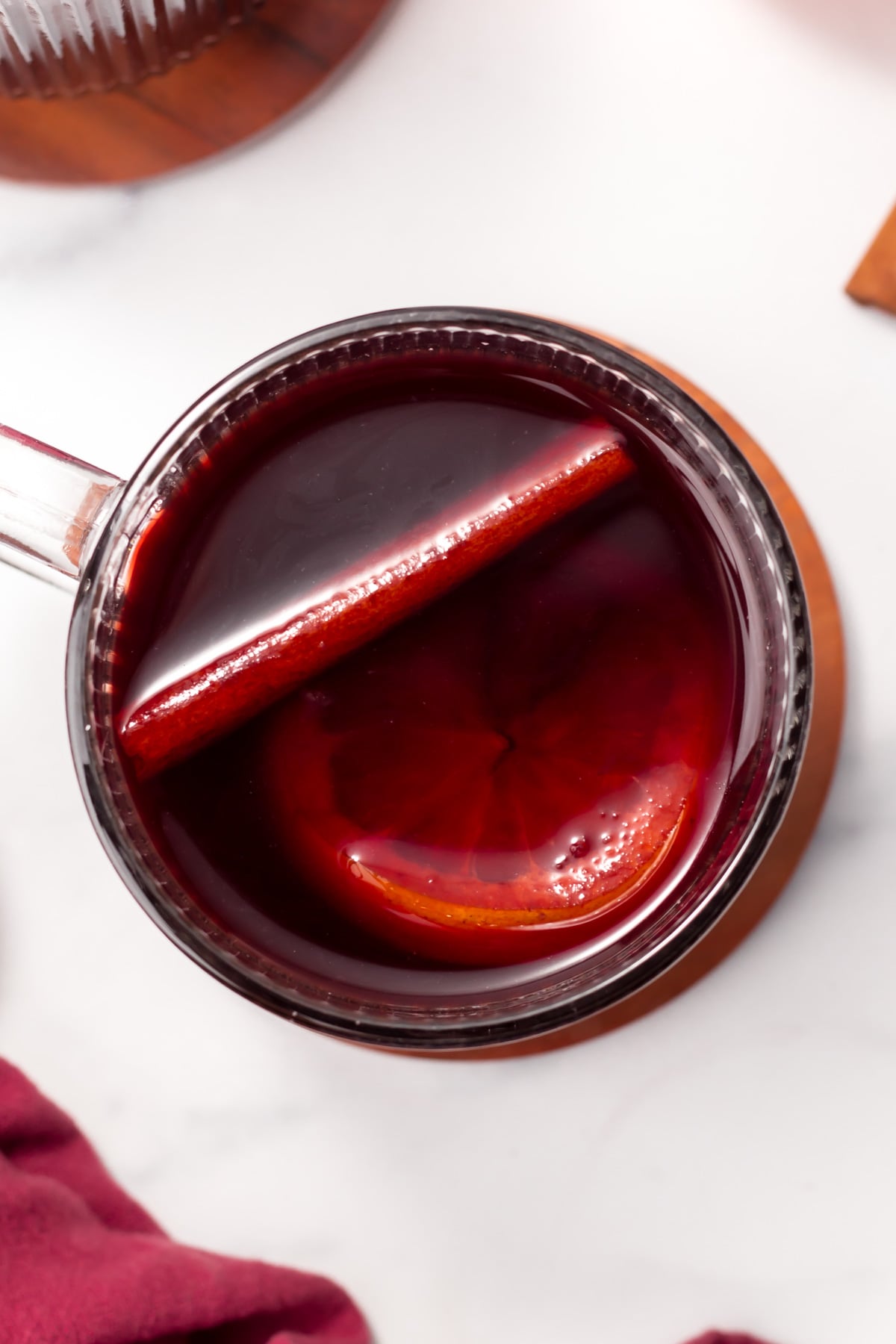 A clear glass mug filled with dark red liquid, containing a slice of orange and a stick of cinnamon, viewed from above on a light surface.
