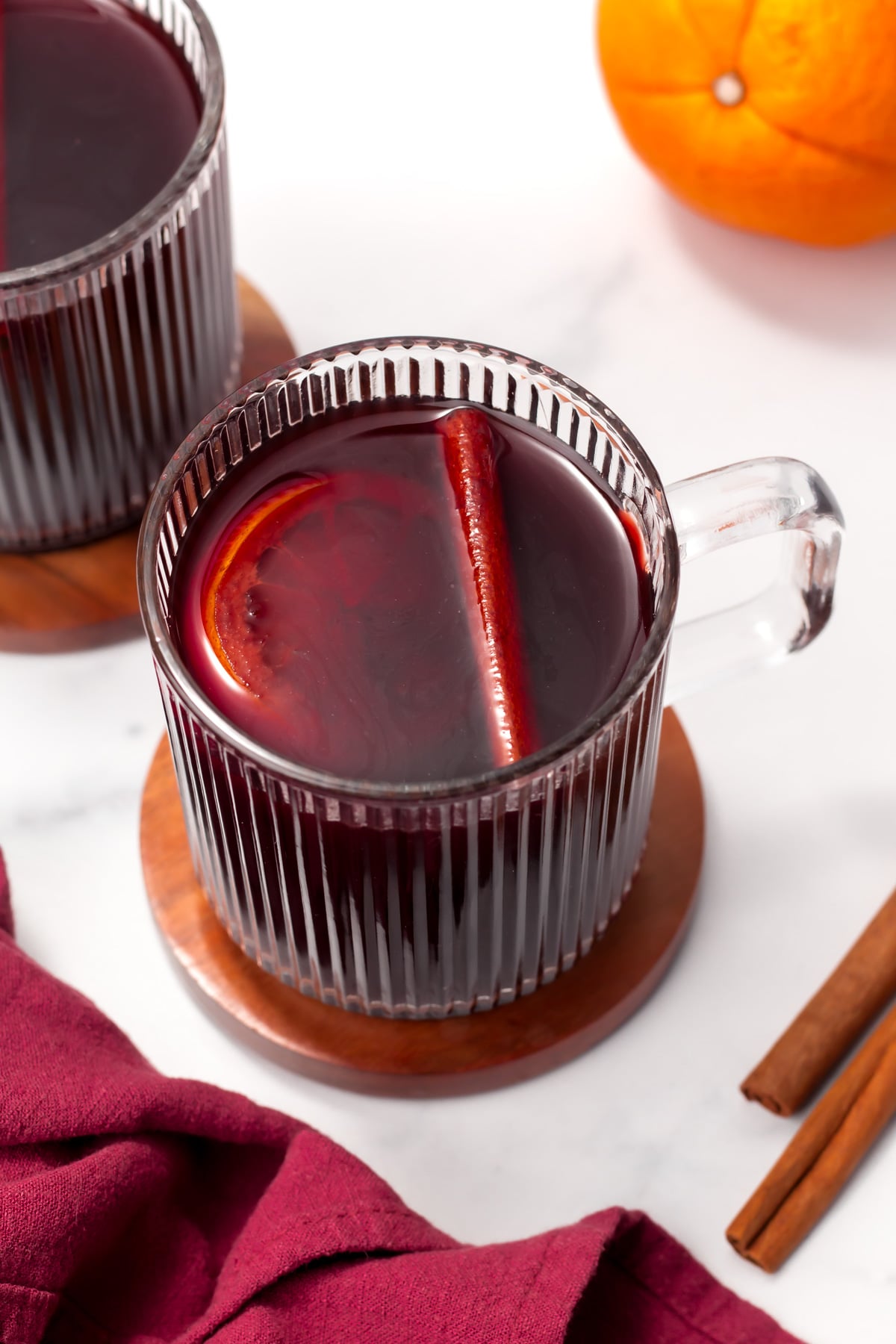 A clear glass mug filled with red mulled wine, garnished with a slice of orange and a cinnamon stick, sits on a wooden coaster next to a red napkin and cinnamon sticks.
