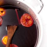 A pot filled with dark red liquid, orange slices, a cinnamon stick, and a wooden spoon, sitting on a white surface.