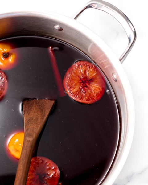 A pot filled with dark red liquid, orange slices, a cinnamon stick, and a wooden spoon, sitting on a white surface.