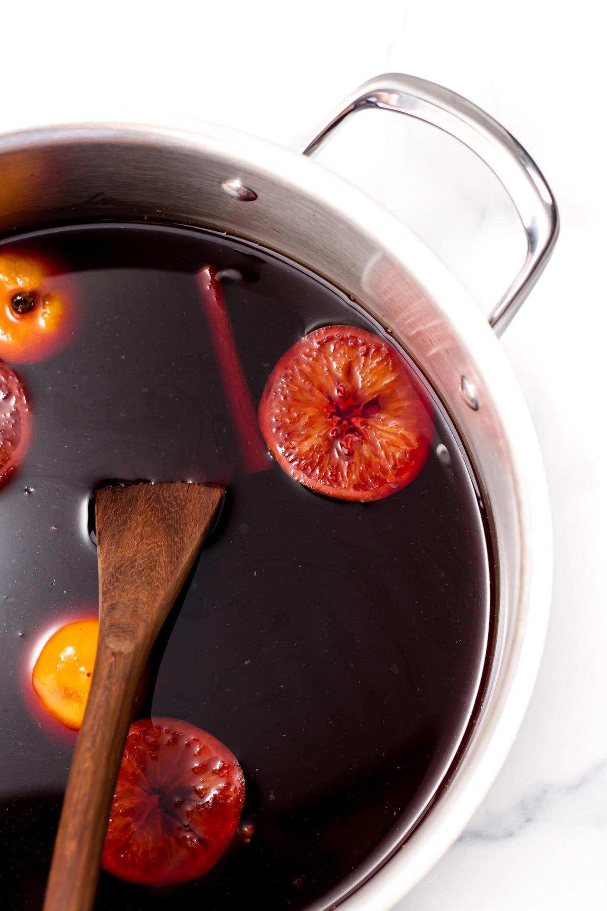 A pot filled with dark red liquid, orange slices, a cinnamon stick, and a wooden spoon, sitting on a white surface.