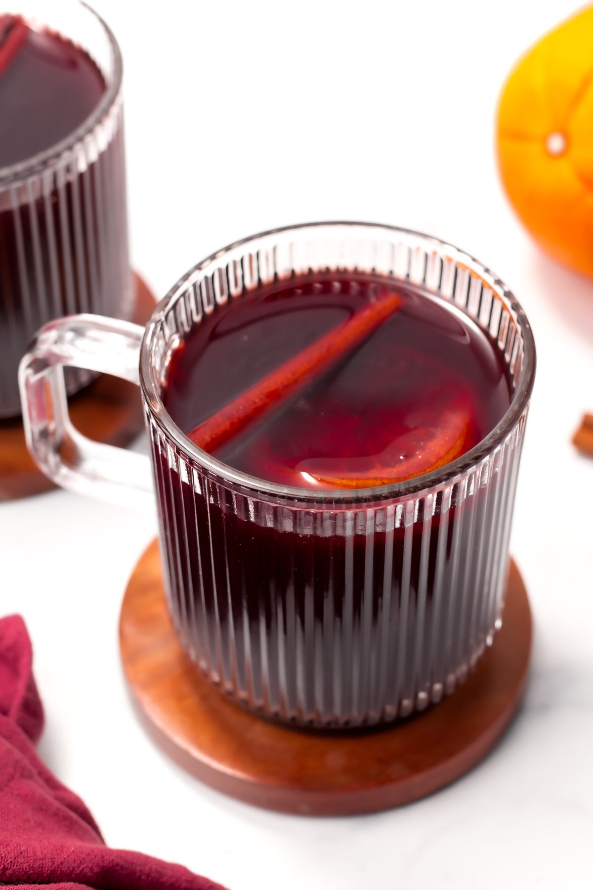 A clear glass mug filled with dark red mulled wine, garnished with a cinnamon stick and an orange slice, sits on a wooden coaster.