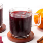A clear glass mug filled with dark red liquid sits on a wooden coaster beside an orange and cinnamon sticks.