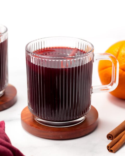 A clear glass mug filled with dark red liquid sits on a wooden coaster beside an orange and cinnamon sticks.