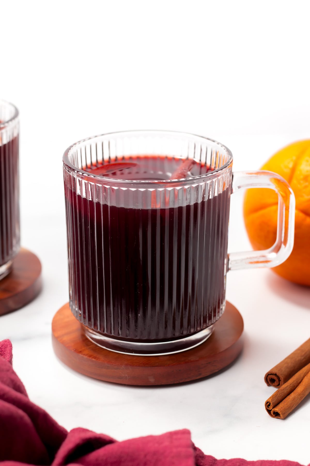 A clear glass mug filled with dark red liquid sits on a wooden coaster beside an orange and cinnamon sticks.