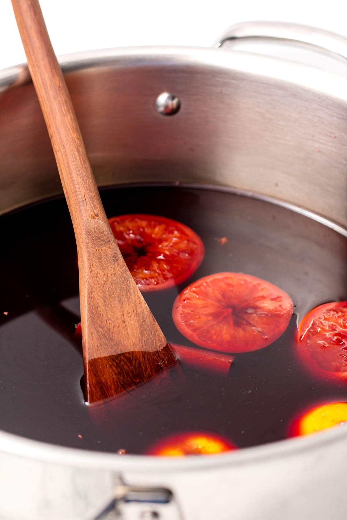 A stainless steel pot filled with dark liquid and orange slices, stirred with a wooden spoon.