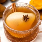 A glass cup of spiced tea garnished with a pear slice, cinnamon stick, and star anise, on a white surface with spices nearby.