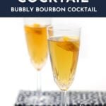 Two champagne flutes filled with a light amber Seelbach bourbon cocktail, each garnished with an orange twist, are set on a white surface with a patterned cloth nearby.
