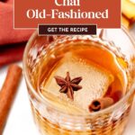 A glass of chai old-fashioned with a large ice cube, garnished with star anise and a cinnamon stick. Text overlay reads: "Chai Old-Fashioned—Get the Recipe.