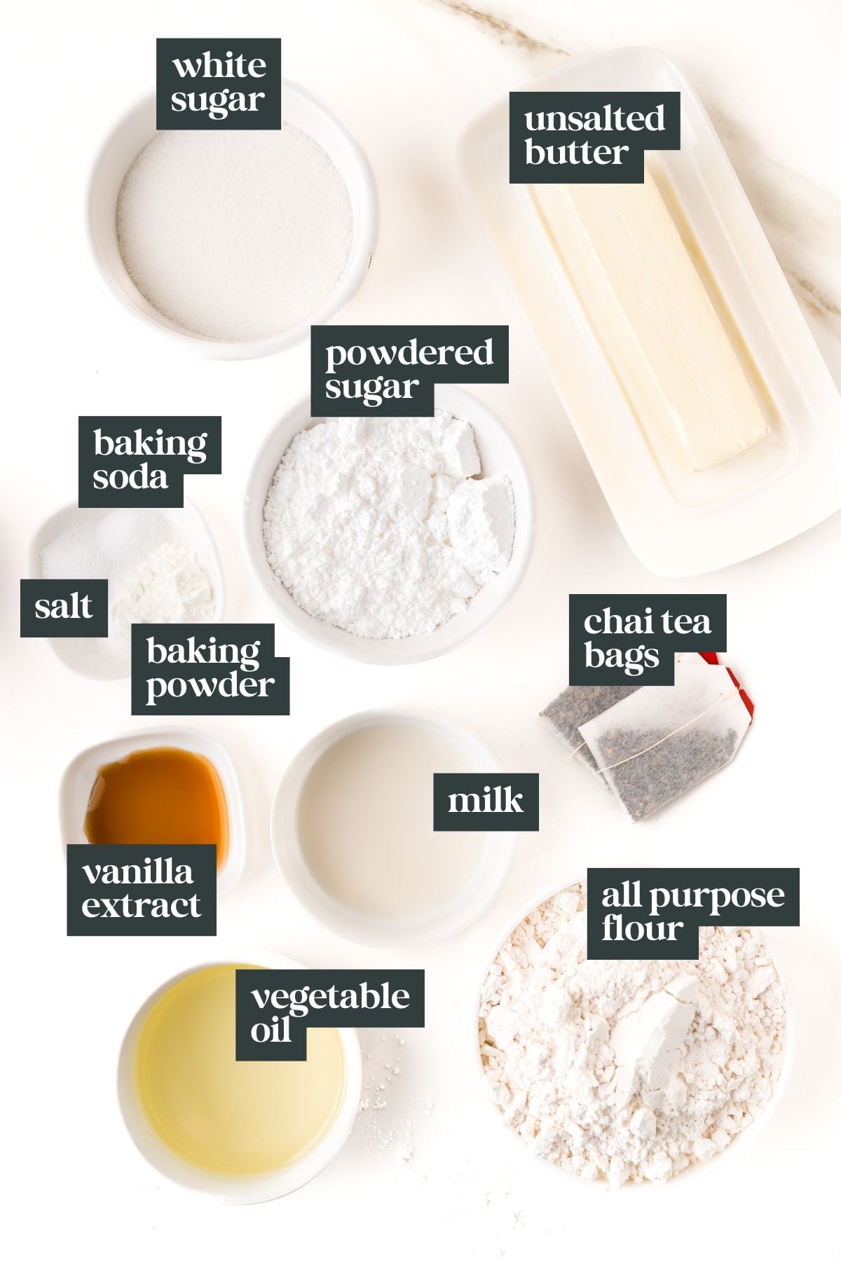 Various baking ingredients and their labels, perfect for chai sugar cookies, including white sugar, unsalted butter, powdered sugar, baking soda, baking powder, salt, chai tea bags, vanilla extract, milk, vegetable oil, and flour.