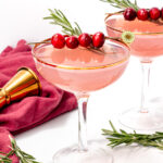 Two pink cocktails in coupe glasses, garnished with cranberries and rosemary sprigs, are set on a white surface with a red cloth and a gold jigger nearby.