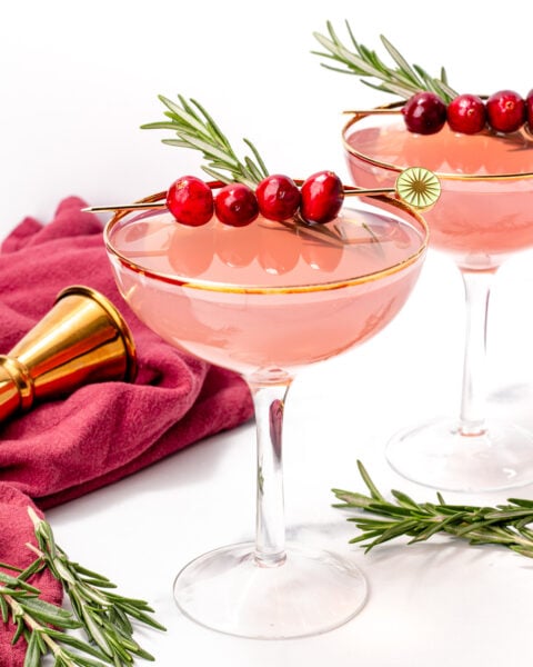 Two pink cocktails in coupe glasses, garnished with cranberries and rosemary sprigs, are set on a white surface with a red cloth and a gold jigger nearby.