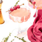 Two pink cocktails in scalloped glasses, garnished with sugared cranberries and thyme sprigs, with a gold jigger and burgundy cloth nearby on a white surface.