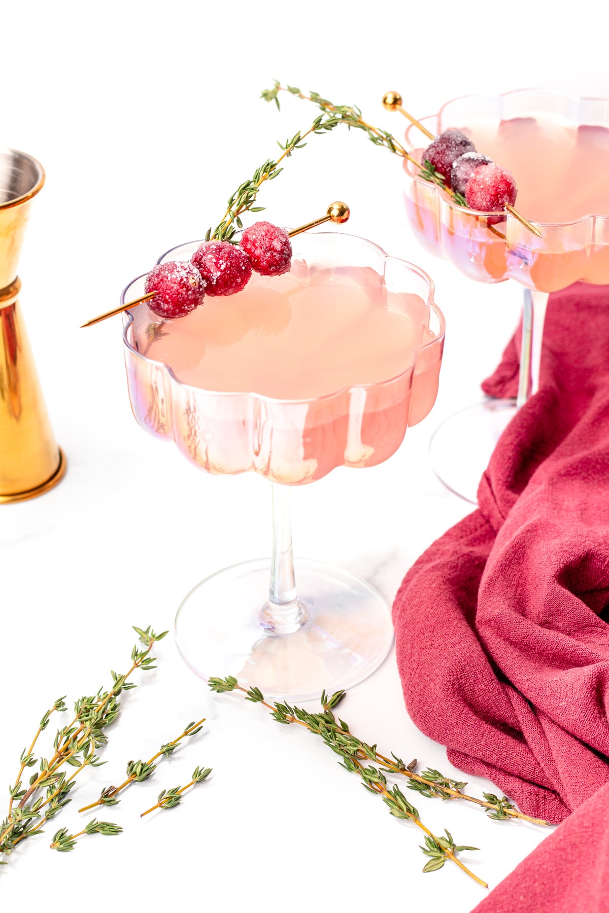 Two pink cocktails in scalloped glasses, garnished with sugared cranberries and thyme sprigs, with a gold jigger and burgundy cloth nearby on a white surface.