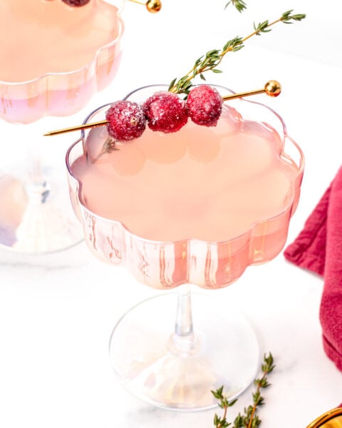 A pink cocktail in a scalloped glass, garnished with sugared cranberries and a sprig of thyme on a cocktail pick, with a red napkin and thyme sprigs nearby.