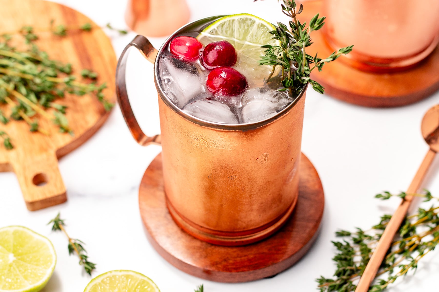 A copper mug filled with ice, garnished with lime, cranberries, and thyme, sits on a wooden coaster surrounded by herbs and lime halves.