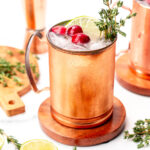 A copper mug filled with ice, garnished with cranberries, a lime slice, and thyme, sits on a wooden coaster; bar tools and lime halves are nearby.