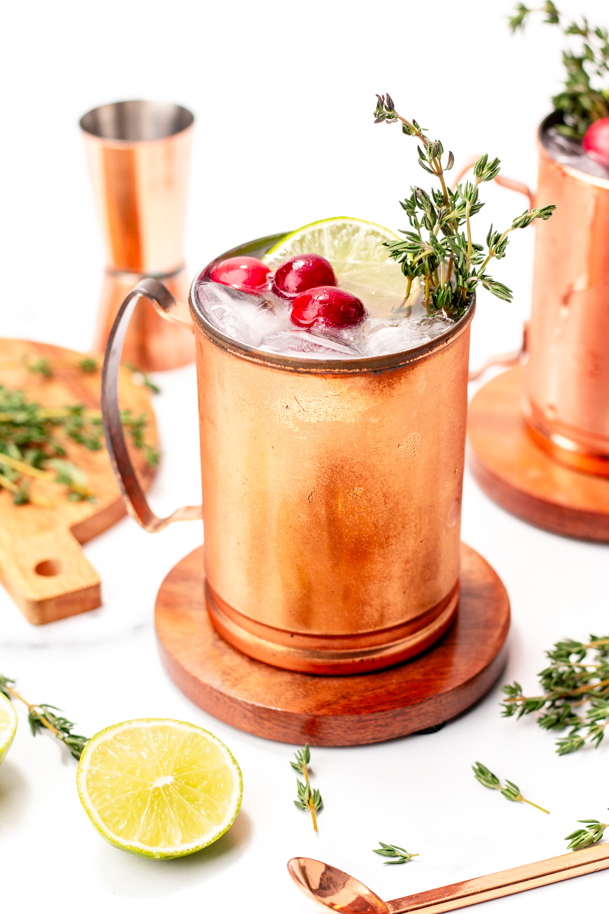 A copper mug filled with ice, garnished with cranberries, a lime slice, and thyme, sits on a wooden coaster; bar tools and lime halves are nearby.