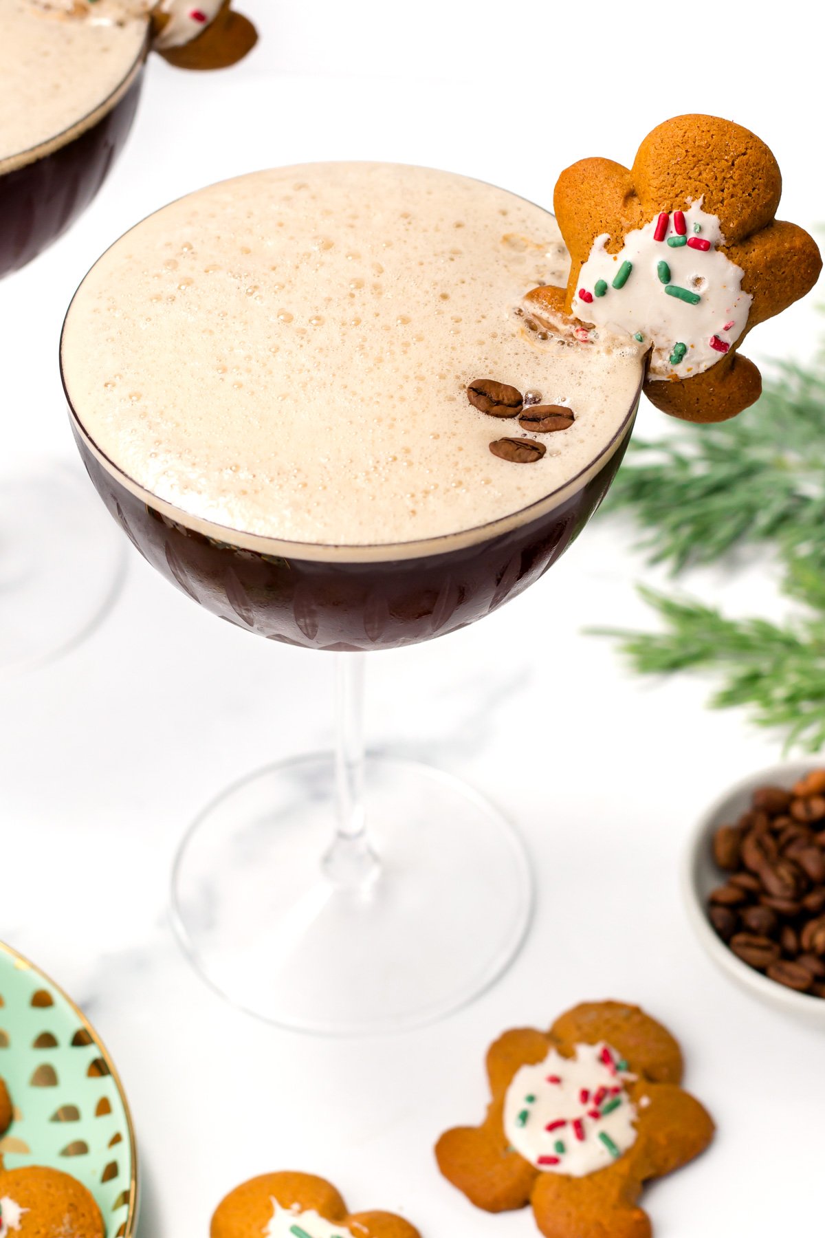 A cocktail in a coupe glass topped with foam, garnished with coffee beans and a decorated gingerbread cookie; festive decorations and cookies nearby.