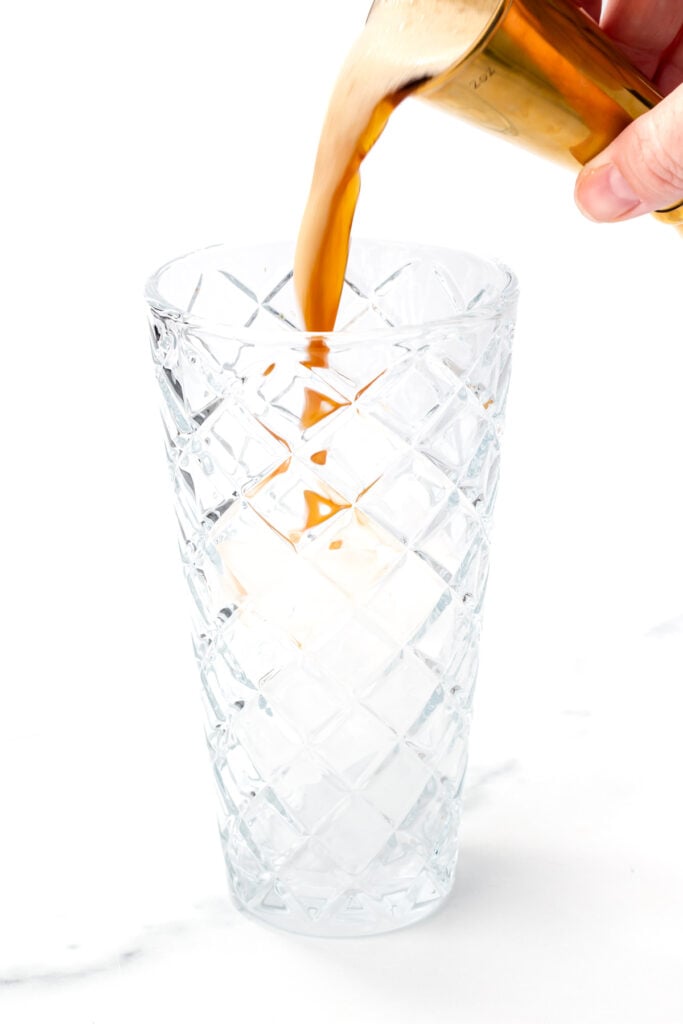 A hand pours a brown liquid from a metal cup into a tall, empty, textured glass on a white background.