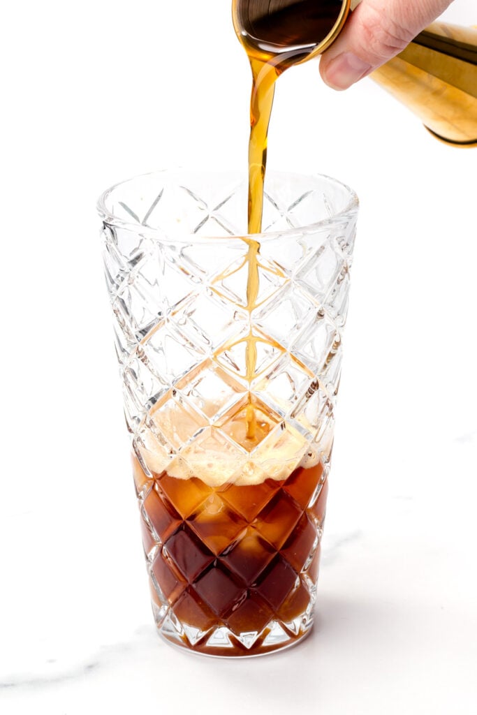 Iced coffee is being poured from a metal container into a patterned glass filled with ice cubes on a white background.
