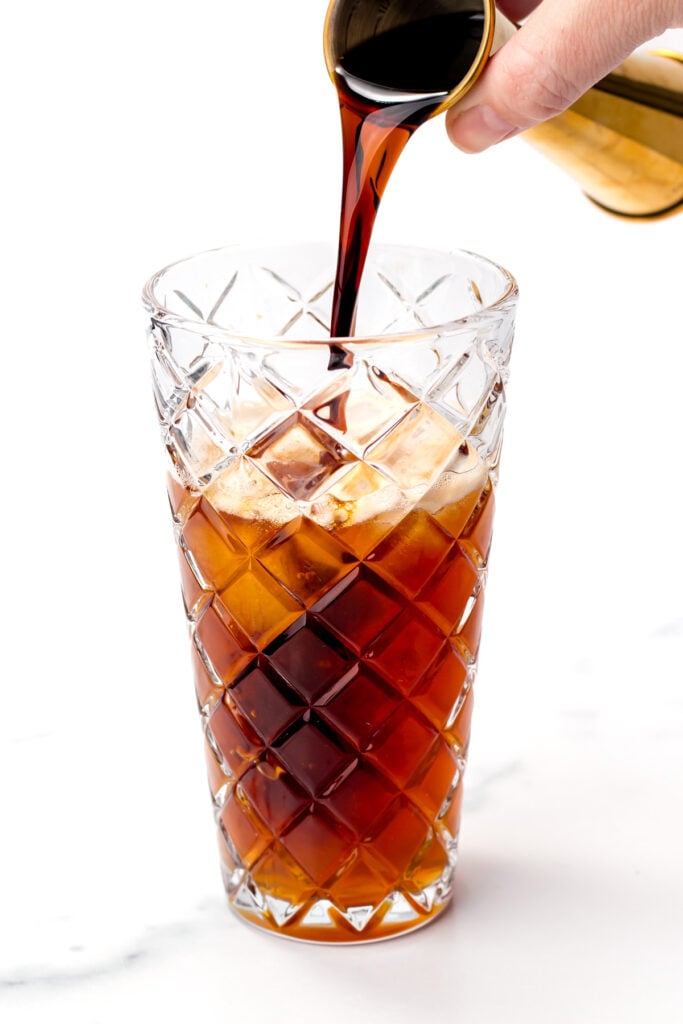 Iced coffee being poured into a clear, patterned glass filled with ice, against a white background.