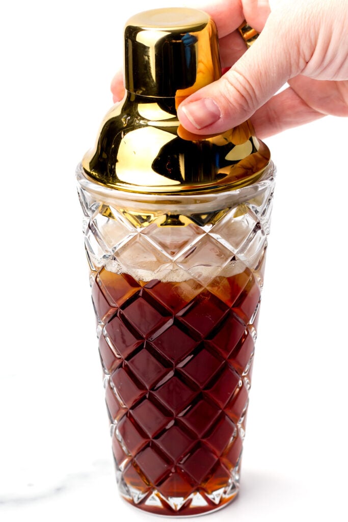A hand holds the lid of a glass cocktail shaker with a gold top, filled with a dark liquid.