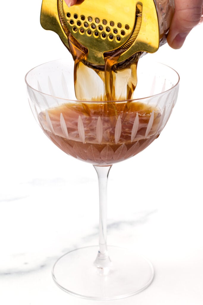 A brown cocktail is being strained from a mixing glass into a decorative coupe glass using a metal strainer.