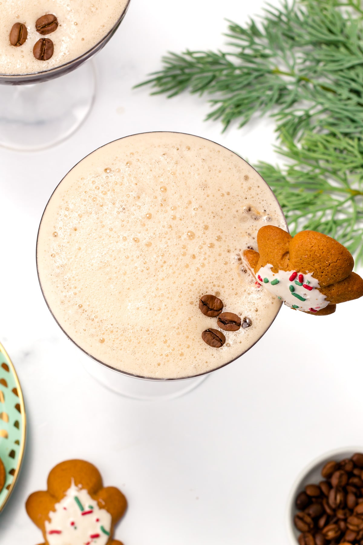 A foamy coffee drink in a glass garnished with coffee beans and a decorated gingerbread cookie, with a sprig of greenery and coffee beans nearby.