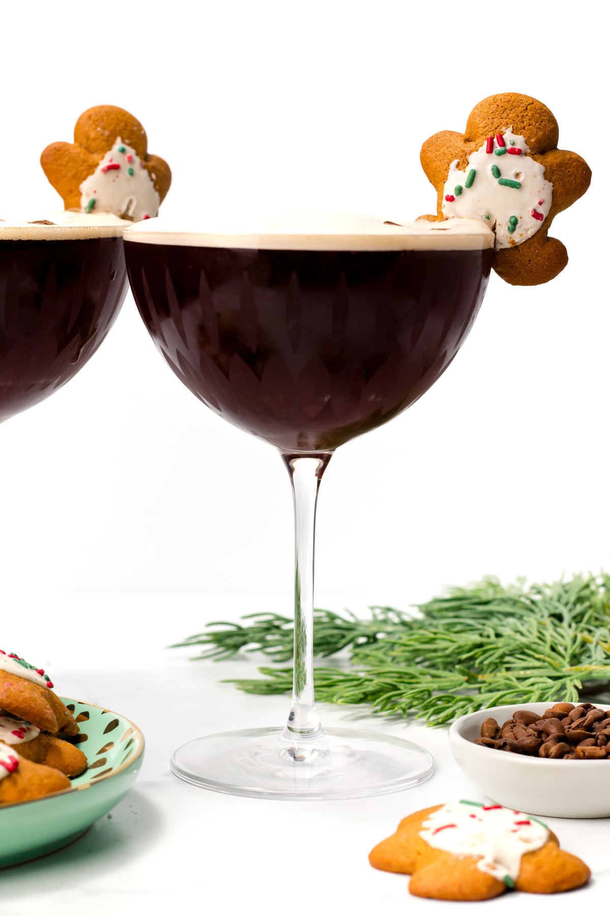 A glass of dark cocktail garnished with a decorated gingerbread cookie, surrounded by coffee beans, greenery, and more cookies.