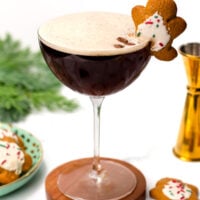 A cocktail in a coupe glass topped with foam, garnished with a gingerbread cookie and coffee beans, sits on a wooden coaster with decorations and cookies nearby.