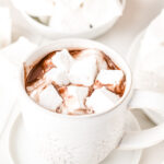 A white mug filled with hot chocolate topped with large marshmallows, surrounded by more marshmallows on white plates.