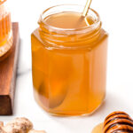 A hexagonal glass jar filled with honey-like liquid, a golden spoon inside, fresh ginger root and a honey dipper with honey nearby, all on a white surface.