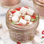A small glass jar filled with hot cocoa mix, topped with mini marshmallows and colorful holiday-themed sprinkles.