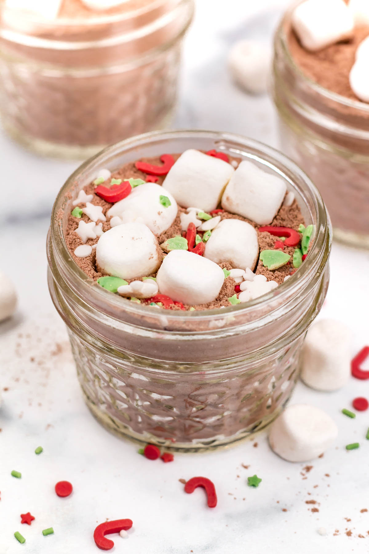 A small glass jar filled with hot cocoa mix, topped with mini marshmallows and colorful holiday-themed sprinkles.