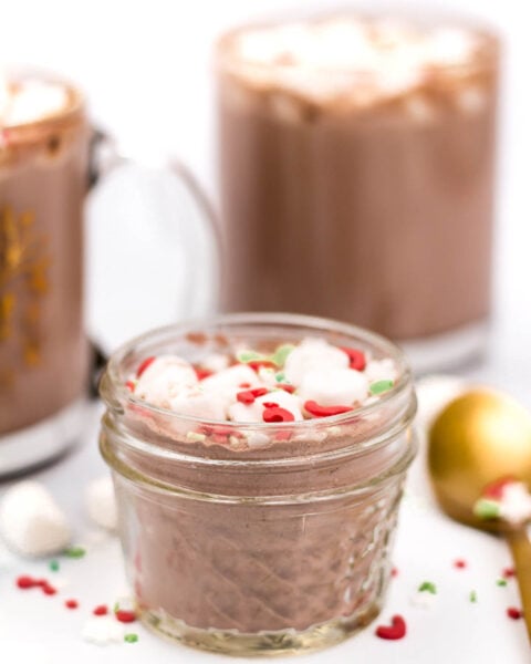 A small glass jar filled with hot chocolate, topped with whipped cream and red and green sprinkles, sits in front of two mugs of hot chocolate.