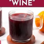 A glass mug filled with dark red non-alcoholic mulled wine sits on a coaster, with part of an orange visible in the background. Text overlays promote a recipe from feastandwest.com.