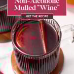 A clear glass mug filled with non-alcoholic mulled "wine," garnished with an orange slice and cinnamon stick, sits on a wooden coaster next to another mug on a white surface.