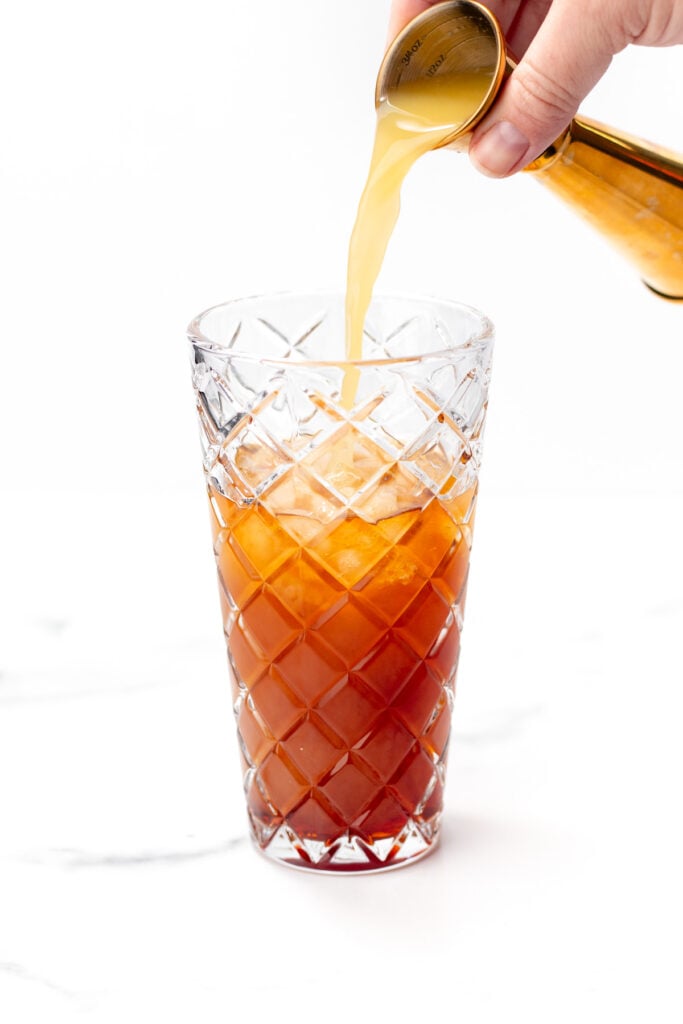 A hand pours a light-colored liquid from a jigger into a tall glass filled with an amber beverage and ice, against a plain white background.