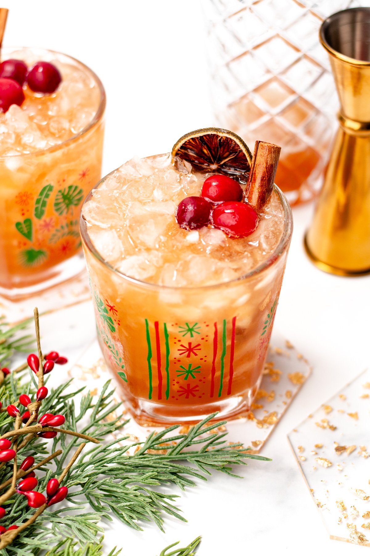 Two festive cocktails with crushed ice, cranberries, a cinnamon stick, and a dried citrus slice, in decorative glasses on coasters, with a gold jigger and greenery nearby.