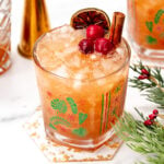 A festive cocktail with crushed ice, garnished with cranberries, a dried citrus slice, and a cinnamon stick, served in a decorated glass on a holiday-themed coaster.
