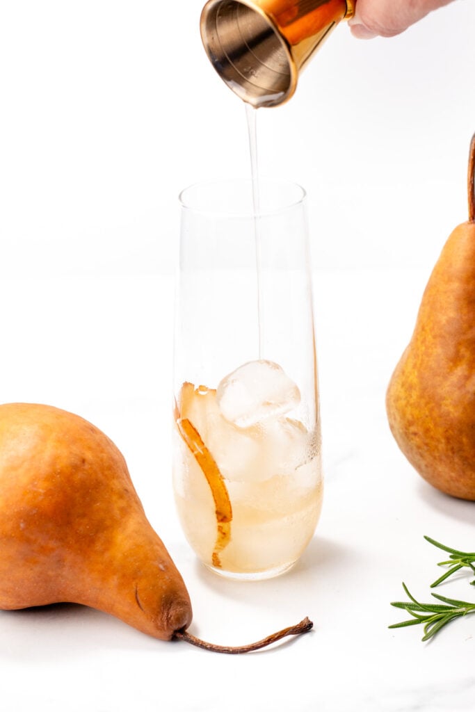 A hand pours liquid from a gold jigger into a glass with ice and a pear slice, surrounded by two whole pears and a sprig of rosemary.