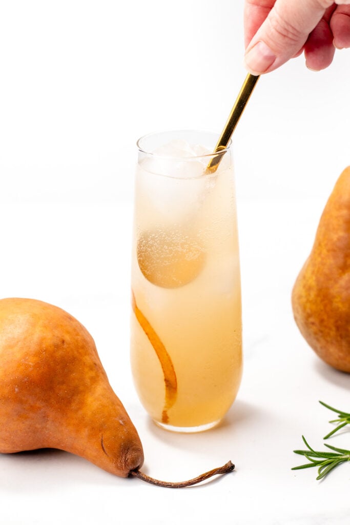 A hand stirs a light brown drink with ice and pear slices in a tall glass, placed between two whole pears on a white surface.