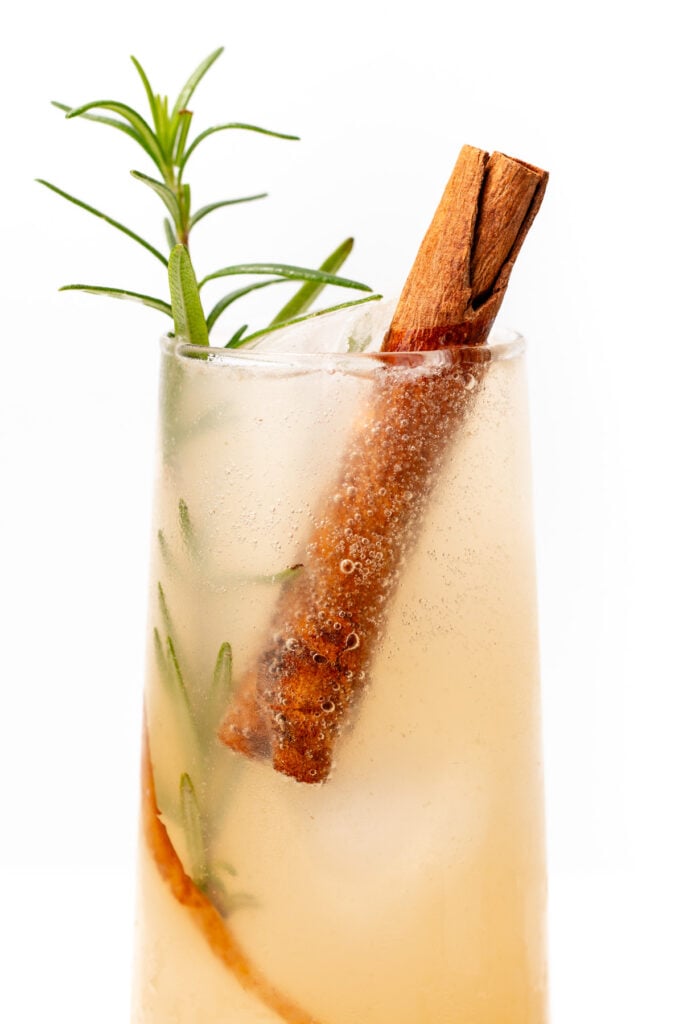 A light-colored cocktail served in a clear glass with ice, garnished with a cinnamon stick and a sprig of rosemary.