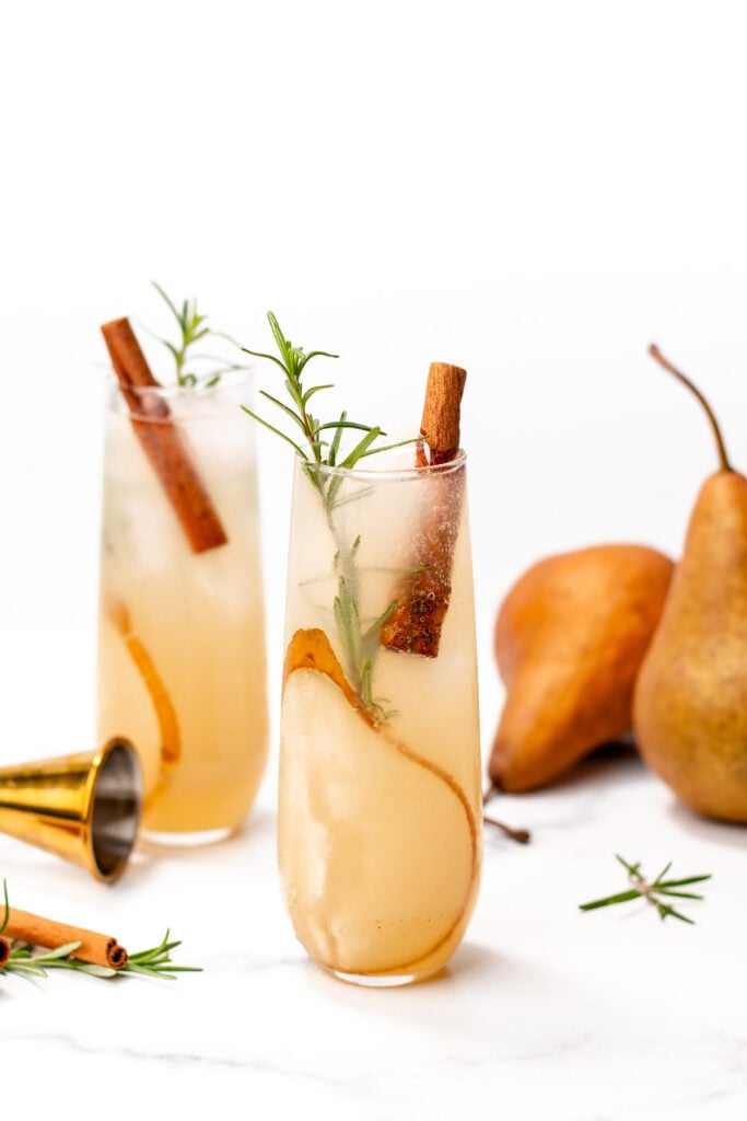 Two glasses of pear cocktails garnished with pear slices, cinnamon sticks, and rosemary sprigs, with whole pears and a jigger in the background on a white surface.