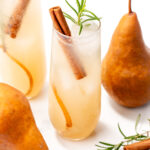 Two glasses of pear-colored drinks with ice, cinnamon sticks, and rosemary sprigs, surrounded by fresh pears, cinnamon sticks, and rosemary on a white background.