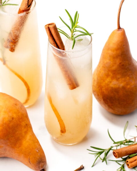 Two glasses of pear-colored drinks with ice, cinnamon sticks, and rosemary sprigs, surrounded by fresh pears, cinnamon sticks, and rosemary on a white background.