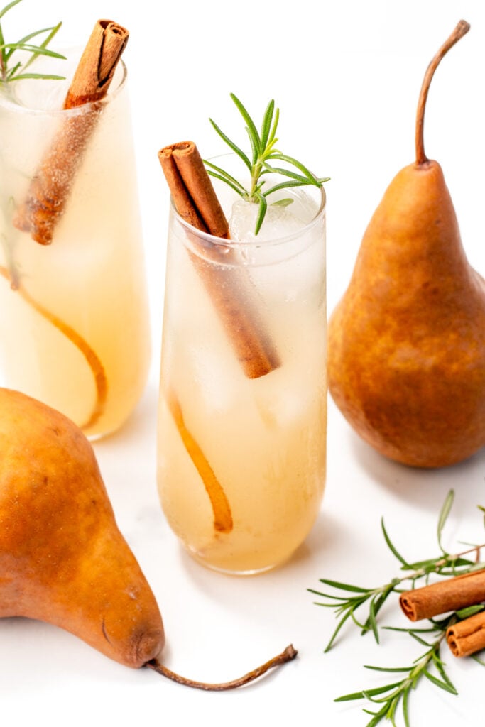 Two glasses of pear-colored drinks with ice, cinnamon sticks, and rosemary sprigs, surrounded by fresh pears, cinnamon sticks, and rosemary on a white background.