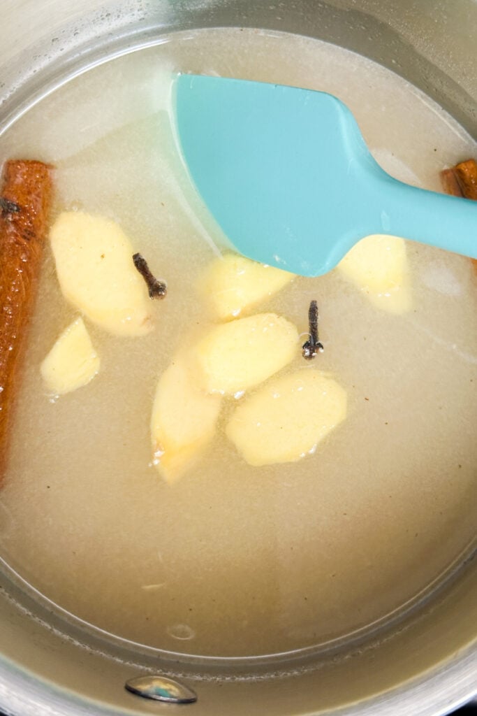 A saucepan with liquid for non-alcoholic mulled wine, sliced ginger, cinnamon stick, and whole cloves being stirred with a blue spatula.