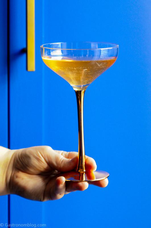 A hand holding a gold-stemmed cocktail glass filled with a light brown beverage in front of a bright blue background.